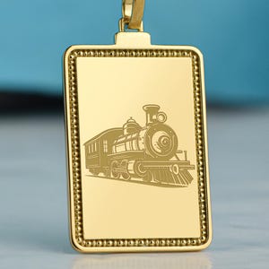May include: A gold-toned rectangular pendant necklace featuring a detailed engraving of a steam locomotive train. The pendant has a beaded border and is suspended from a matching gold-toned rope chain, creating a cohesive metallic accessory.