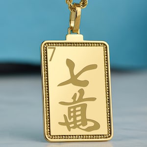 May include: Gold-colored pendant necklace with a rectangular pendant. The pendant features Chinese characters and a beaded border. The necklace chain is a rope style. The background is a light blue color.