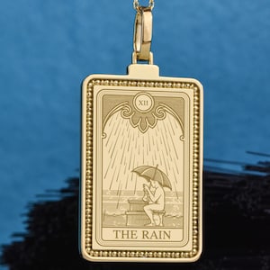 Könnte beinhalten: Ein goldener rechteckiger Anhänger an einer feinen Kette mit einer eingravierten Tarotkarten-Abbildung. Das Motiv zeigt eine Figur unter einem Regenschirm, darüber die römische Ziffer XII und am unteren Rand der Schriftzug THE RAIN.