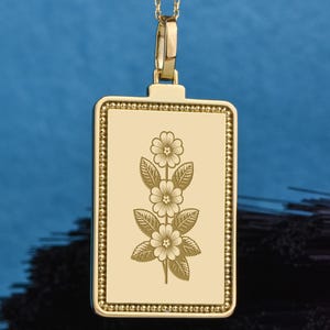 May include: A gold-colored pendant necklace with a rectangular pendant. The pendant features an engraved floral design with three flowers and leaves. The pendant is suspended from a delicate gold chain with small ball accents.