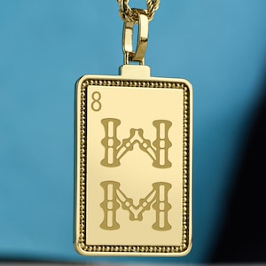 Könnte beinhalten: Goldfarbene Anhängerhalskette mit rechteckigem Anhänger. Der Anhänger zeigt die Zahl "8" und zwei stilisierte "M"-Symbole. Die Halskette hat eine Seilkette und einen Verschluss. Der Hintergrund ist einfarbig blau.