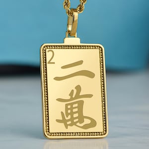 May include: Gold-toned rectangular pendant necklace with a rope chain. The pendant features Chinese characters and the number 2. The pendant has a beaded border and is suspended from a gold-colored chain.