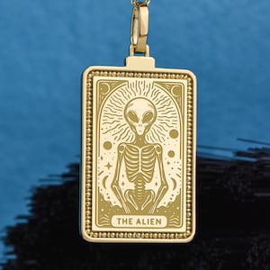 Pode incluir: Um pingente retangular de tom dourado em uma corrente, apresentando uma ilustração gravada de uma figura extraterrestre com um torso esquelético. O design inclui motivos celestiais como estrelas e círculos, com o texto THE ALIEN.