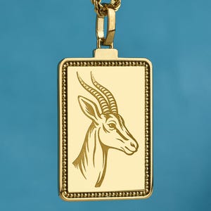 May include: A gold-toned rectangular pendant necklace featuring a detailed engraving of an antelope head in profile. The pendant has a beaded border and is suspended from a matching gold-toned rope chain, creating a cohesive metallic look.