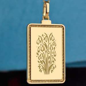 May include: Gold-colored pendant necklace with a rectangular pendant featuring an engraved floral design. The pendant hangs from a delicate chain with small, round beads. The background is a solid blue.