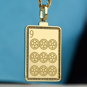 May include: Gold-toned pendant necklace with a rectangular charm. The charm features the number "9" at the top and nine circular designs below. The necklace has a rope chain and a clasp. The background is a solid blue.