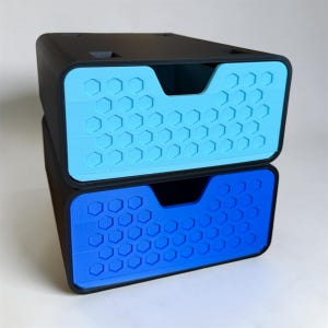 May include: A stack of two black storage drawers with a hexagonal honeycomb pattern on the front panels. The top drawer features a light blue front, while the bottom drawer has a vibrant blue front. Each drawer has a central cutout handle.