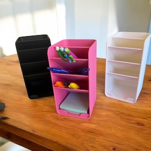 May include: Three desk organizers in black, pink, and white sit on a wooden surface. The pink organizer holds pens, markers, and sticky notes. The black and white organizers have multiple compartments for office supplies.