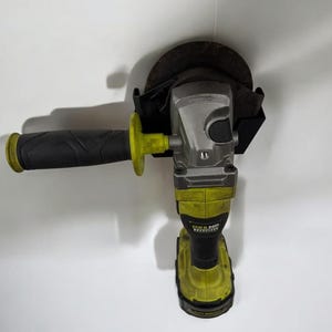May include: A yellow and black cordless angle grinder with a black handle and a black and brown grinding disc. The grinder is mounted on a black wall mount.