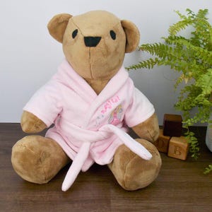 May include: A brown teddy bear wearing a pink robe with a white sash and embroidered with the words "Magical" and a fairy design.