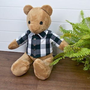 Peut inclure: Un ours en peluche brun portant une chemise à carreaux noirs et blancs avec des boutons bruns. L'ours est assis sur un sol en bois brun.