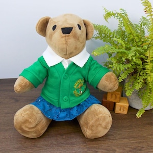 Peut inclure: Un ours en peluche brun portant un cardigan vert avec un logo d'école, une chemise blanche à col et une jupe bleue. Le logo dit "Arden" et a une trèfle vert.