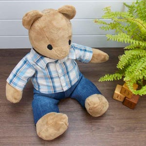 May include: A soft toy teddy bear dressed in a blue and white checked shirt and denim jeans. The bear is light brown with black eyes. Wooden blocks with letters are in the background.