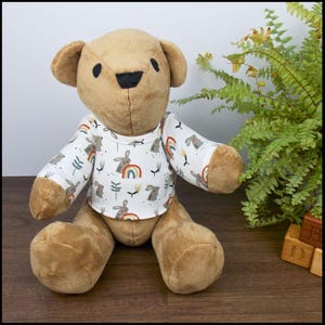 May include: A brown teddy bear wearing a white long-sleeved shirt with a pattern of rainbows and bunnies. The shirt has a crew neck and long sleeves.