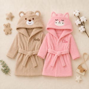 May include: Two plush fleece bathrobes with hoods. The tan robe features a bear face on the hood and the name Henty embroidered in brown. The pink robe features a cat face on the hood and the name Cloe embroidered in pink. Both have tie belts.