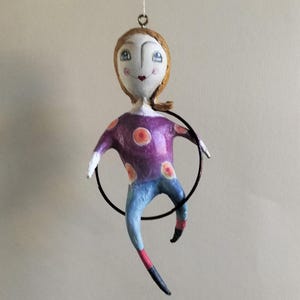 Puede incluir: Adorno de muñeca de papel hecho a mano. La muñeca sonriente tiene cabello castaño y viste una camisa morada con lunares naranjas, vaqueros azules y un aro negro. Una decoración artesanal y original.