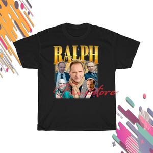 May include: Black t-shirt with a gold-toned graphic of Ralph Fiennes and the name "RALPH". The design incorporates several images of the actor in a vintage style. The shirt has a crew neck.