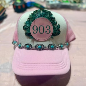 May include: A light pink trucker hat featuring a white front panel with a circular patch displaying the number 903 in green glitter. The patch is surrounded by dark green feathers, and a silver-toned chain with turquoise stones accents the brim.