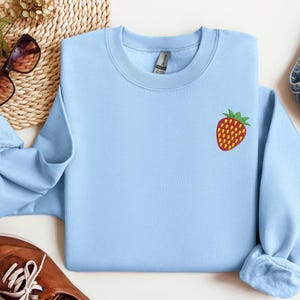 May include: A light blue sweatshirt with a small embroidered strawberry on the chest. The strawberry is red with yellow seeds and green leaves. The sweatshirt is displayed with sunglasses, a woven mat, jeans, and brown shoes.