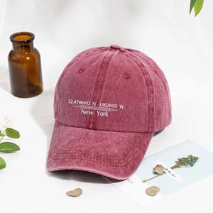 May include: A maroon baseball cap with the text "52.4796992 N -1.9026911 W New York" in white. The cap has a curved brim and a slightly faded appearance. A brown glass bottle is in the background.