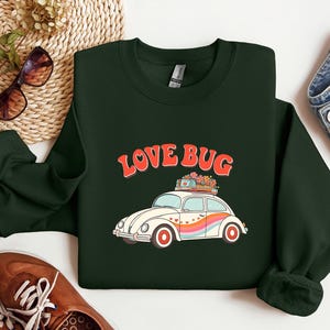 Pode incluir: Camisola verde escuro com o texto "LOVE BUG" acima de uma ilustração de carro retro. O carro é branco com uma faixa de arco-íris e flores no topo. A camisola é mostrada com jeans e sapatos castanhos.