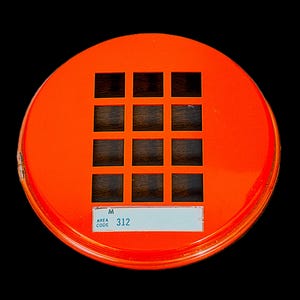 May include: A bright orange, circular object with a grid of square cutouts. The object has a label that reads "AREA CODE 312" and the letter "M". The background is solid black.