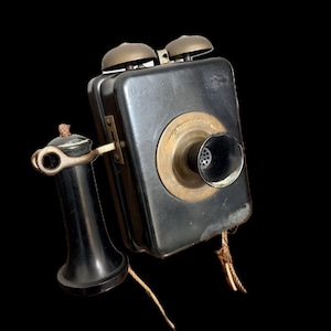 May include: An antique black wall-mounted telephone with a brass speaker and two bells. The handset is black and connected by a fabric-covered cord. The phone is a vintage item.