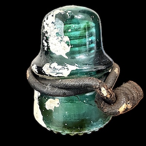 Könnte beinhalten: Ein Vintage-Glasisolator in Teal-Farbe mit dunklem, verwittertem Aussehen. Der Isolator ist mit einem dicken, dunklen, seilartigen Material umwickelt. Weiße Farbe blättert vom Glas ab und verstärkt die gealterte Ästhetik. Der Hintergrund ist tiefschwarz.