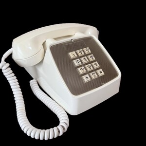May include: A vintage white push-button telephone with a coiled handset cord. The phone has a rectangular base with a keypad featuring numbered buttons and a clear plastic cover. The phone is set against a black background.