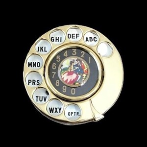 Puede incluir: Un dial de teléfono rotatorio de color dorado con un centro negro que presenta una imagen colorida de una pareja bailando. El dial tiene botones y números con letras blancas, con las letras ABC, DEF, GHI, JKL, MNO, PRS, TUV, WXY y OPTR.