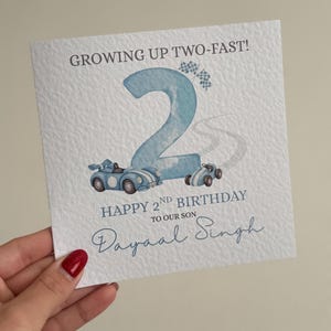 Puede incluir: Una tarjeta de felicitaci&oacute;n cuadrada con superficie blanca texturizada que presenta un n&uacute;mero dos grande en azul claro, dos coches de carreras azules y banderas a cuadros. El texto dice: GROWING UP TWO-FAST! HAPPY 2ND BIRTHDAY TO OUR SON Dayaal Singh.