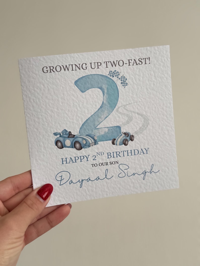 Puede incluir: Una tarjeta de felicitaci&oacute;n cuadrada con superficie blanca texturizada que presenta un n&uacute;mero dos grande en azul claro, dos coches de carreras azules y banderas a cuadros. El texto dice: GROWING UP TWO-FAST! HAPPY 2ND BIRTHDAY TO OUR SON Dayaal Singh.