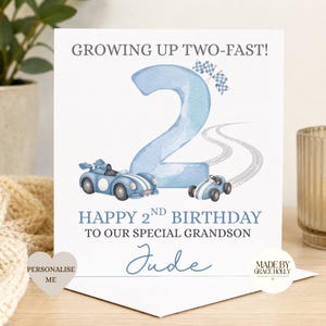 Puede incluir: Una tarjeta de felicitaci&oacute;n cuadrada con un n&uacute;mero dos grande en acuarela azul claro, dos coches de carreras azules y banderas a cuadros. El texto dice: GROWING UP TWO-FAST! HAPPY 2ND BIRTHDAY TO OUR SPECIAL GRANDSON Jude. Incluye huellas.