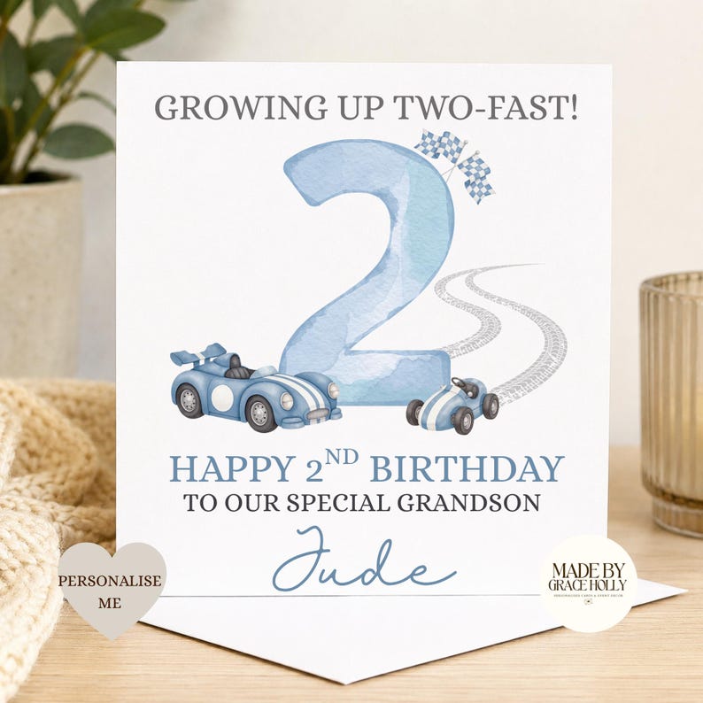 Puede incluir: Una tarjeta de felicitaci&oacute;n cuadrada con un n&uacute;mero dos grande en acuarela azul claro, dos coches de carreras azules y banderas a cuadros. El texto dice: GROWING UP TWO-FAST! HAPPY 2ND BIRTHDAY TO OUR SPECIAL GRANDSON Jude. Incluye huellas.
