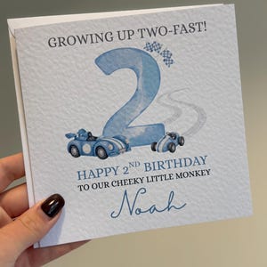 Puede incluir: Una tarjeta de felicitaci&oacute;n cuadrada con superficie blanca texturizada que presenta un n&uacute;mero dos grande en azul claro, dos coches de carreras azules y banderas a cuadros. El texto dice: GROWING UP TWO-FAST! HAPPY 2ND BIRTHDAY TO OUR CHEEKY LITTLE MONKEY y Noah.