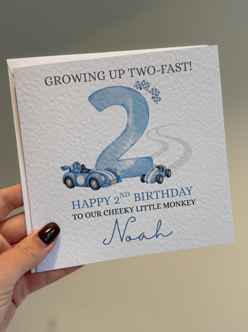 Puede incluir: Una tarjeta de felicitaci&oacute;n cuadrada con superficie blanca texturizada que presenta un n&uacute;mero dos grande en azul claro, dos coches de carreras azules y banderas a cuadros. El texto dice: GROWING UP TWO-FAST! HAPPY 2ND BIRTHDAY TO OUR CHEEKY LITTLE MONKEY y Noah.