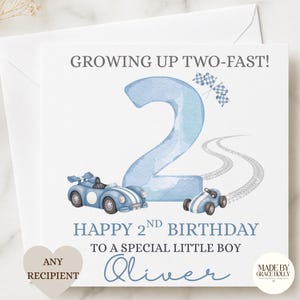 Puede incluir: Una tarjeta de felicitaci&oacute;n cuadrada con un n&uacute;mero dos grande en acuarela azul claro, dos coches de carreras azules y banderas a cuadros. El texto dice: GROWING UP TWO-FAST! HAPPY 2ND BIRTHDAY TO A SPECIAL LITTLE BOY Oliver. Incluye huellas.