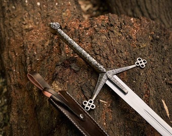 May include: A detailed image of a medieval sword with an ornate silver handle and crossguard. The blade is silver and the sword rests on a textured, brown wooden surface. A brown leather sheath is also visible.
