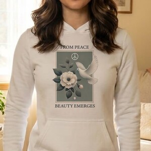 Könnte beinhalten: Weißer Kapuzenpullover mit einem Front-Grafik-Design mit einer Taube, Blumen und dem Text "FROM PEACE" und "BEAUTY EMERGES". Der Hoodie hat eine Kängurutasche und eine Kapuze mit Kordelzug. Das Model trägt Jeansshorts.