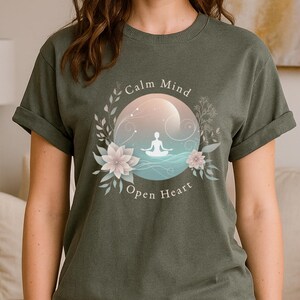 Puede incluir: Camiseta verde musgo con un dise&ntilde;o circular con las palabras "Calm Mind Open Heart". El dise&ntilde;o incluye una persona meditando, rodeada de flores y hojas, con un fondo rosa claro y azul.