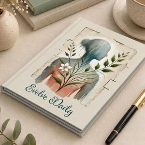 Könnte beinhalten: Ein Notizbuch mit festem Einband in Cremefarben, das eine Aquarellillustration einer menschlichen Silhouette zeigt, die mit grünen Blattzweigen und einer weißen Blume verwoben ist. Der Text Evolve Daily steht in dunkelgrüner Schreibschrift darunter.
