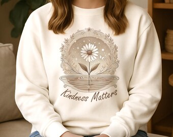 May include: A cream-coloured crewneck sweatshirt featuring a central graphic of a white daisy inside a circular, ornate frame with leaves and stars. Below the flower, a reflection appears in water, accompanied by the text "Kindness Matters" in brown.