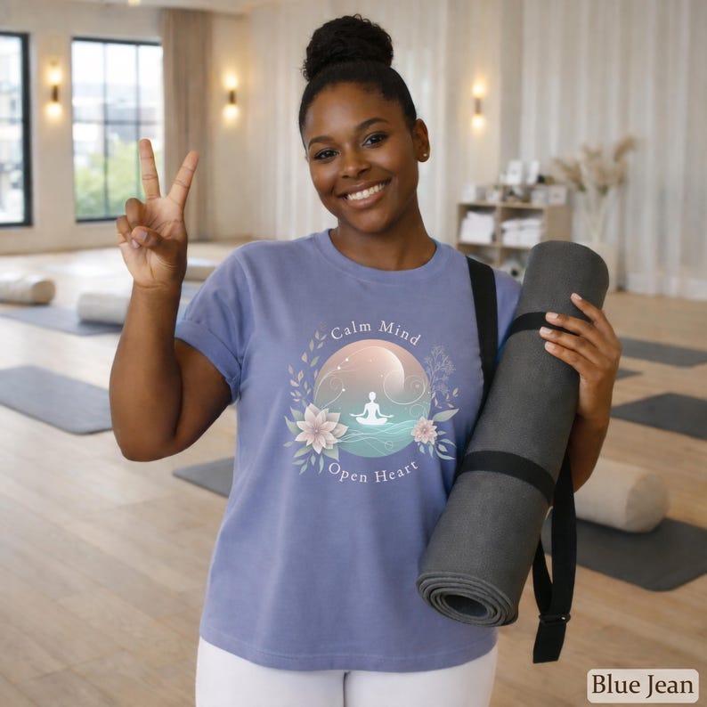 Puede incluir: Una camiseta azul vaquera con las palabras "Calm Mind Open Heart" y un gr&aacute;fico de una persona meditando. La persona sostiene una esterilla de yoga gris enrollada con una correa negra. El fondo es un estudio de yoga.