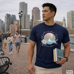Puede incluir: Camiseta azul marino con un dise&ntilde;o circular que presenta una figura meditando, flores y las palabras "Calm Mind Open Heart". El fondo muestra un horizonte urbano y agua.