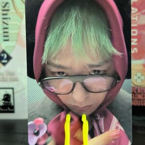 May include: A trading card featuring a person with green hair wearing a pink hoodie and glasses. The card is held by a bright yellow stand. The background includes text from other cards.