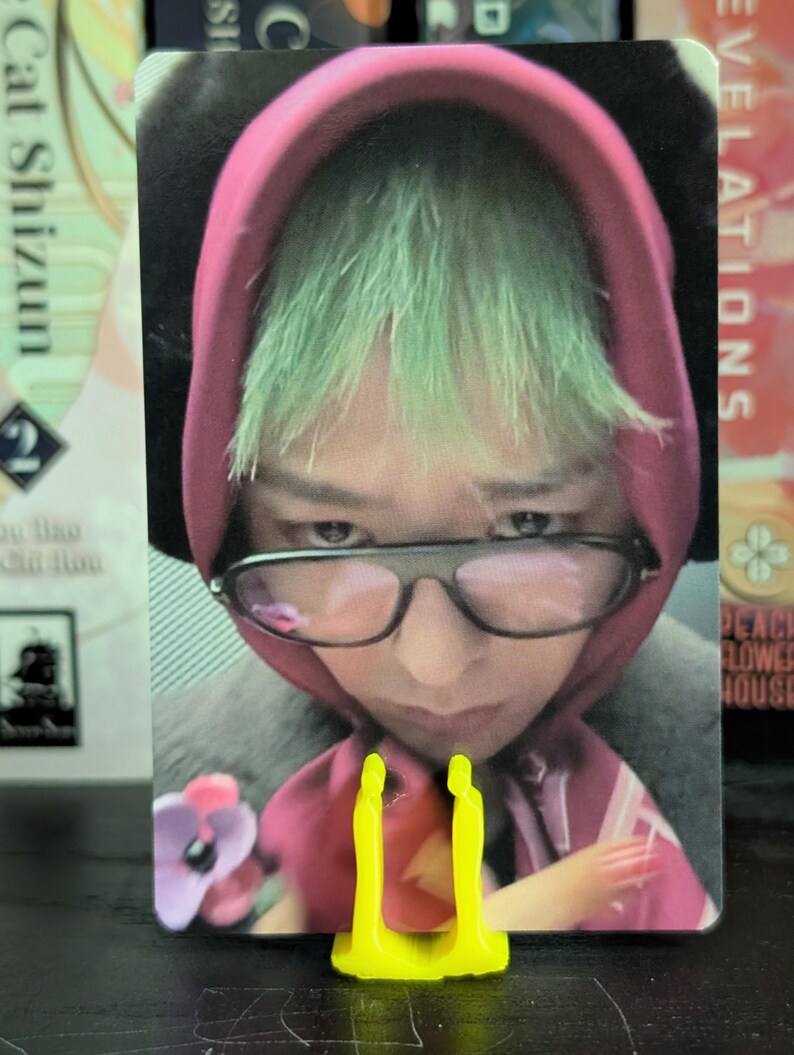 May include: A trading card featuring a person with green hair wearing a pink hoodie and glasses. The card is held by a bright yellow stand. The background includes text from other cards.