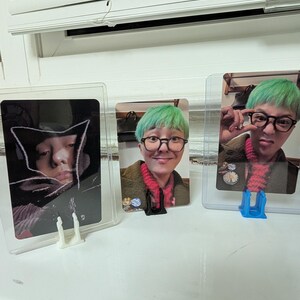 May include: Three photo cards in protective sleeves. The cards feature images of a person with green hair and glasses. The cards are displayed in clear plastic holders on a white surface near a window.