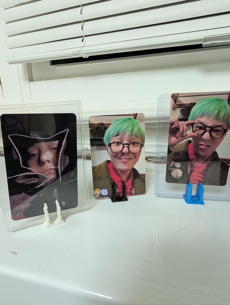 May include: Three photo cards in protective sleeves. The cards feature images of a person with green hair and glasses. The cards are displayed in clear plastic holders on a white surface near a window.