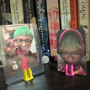 May include: Two trading cards featuring a person with green hair and glasses, displayed in protective sleeves. The cards are propped up by yellow and pink card stands, set against a backdrop of books.