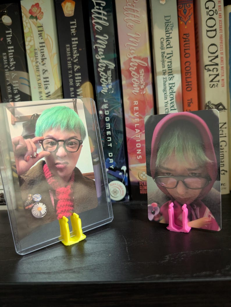 May include: Two trading cards featuring a person with green hair and glasses, displayed in protective sleeves. The cards are propped up by yellow and pink card stands, set against a backdrop of books.